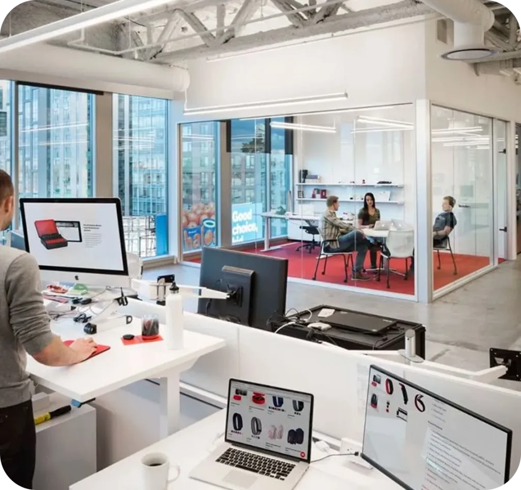 A bright, modern open-plan office with standing desks and desktop computers in the foreground, and a glass-walled meeting room in the background where a small team is collaborating, featuring large windows, natural light, and a clean, contemporary workspace design.