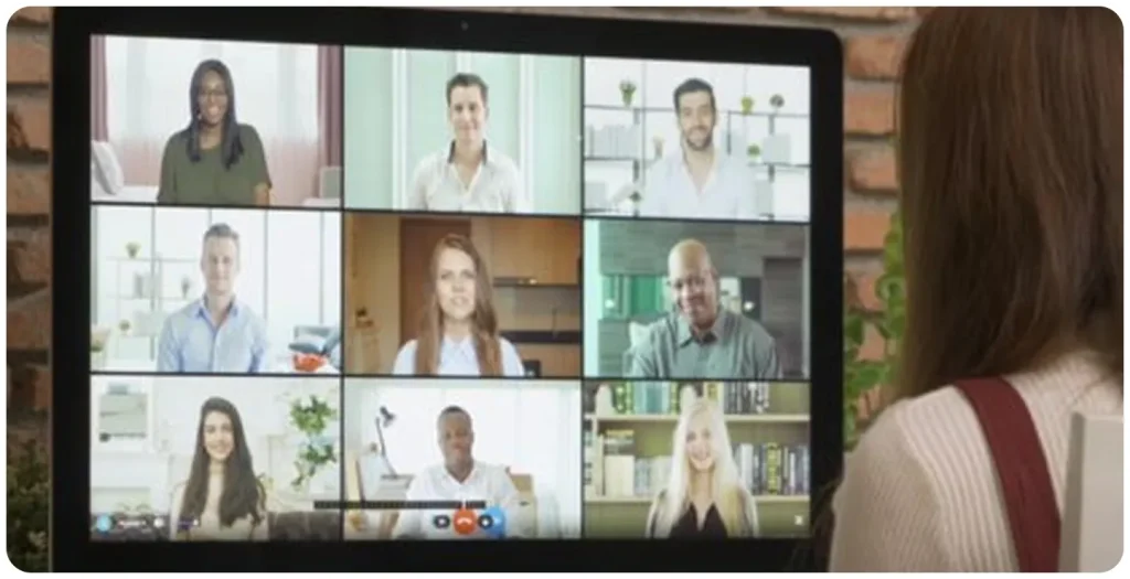 A professional video conference displayed on a large screen, showing a diverse group of nine people in a virtual meeting grid, with a woman viewed from behind attending the online business call in a modern workspace.