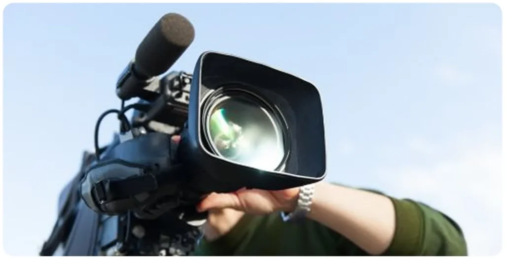 Close-up of a professional video camera with a large lens hood and mounted shotgun microphone, held by a camera operator outdoors under a clear sky, representing professional videography and filming production.