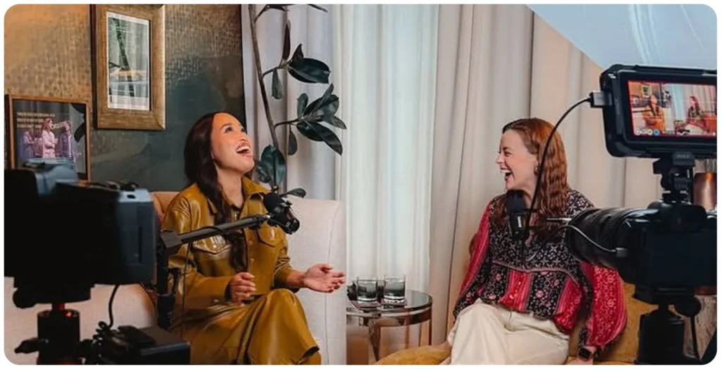 Two women recording a podcast or interview in a cozy indoor studio, sitting on chairs with microphones, cameras, and lighting equipment, smiling and laughing during a professional video and audio content production session.