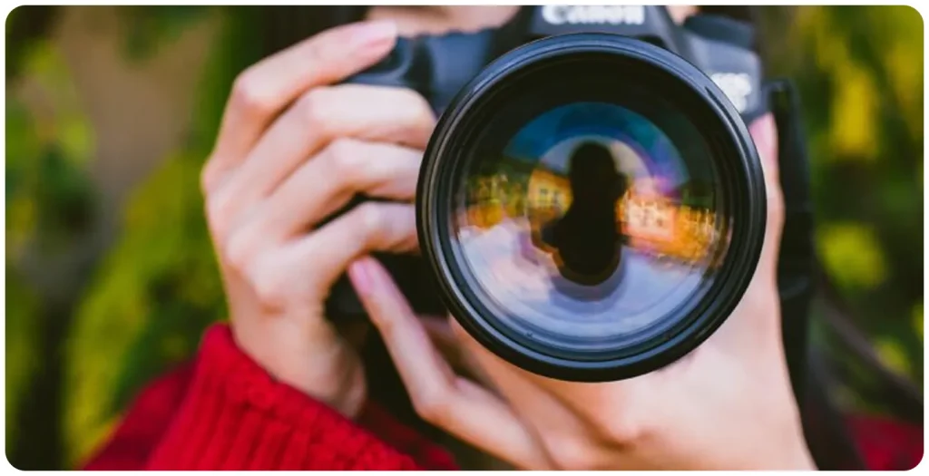 Close-up of a photographer holding a DSLR camera, with the subject reflected in the camera lens against a blurred green outdoor background.