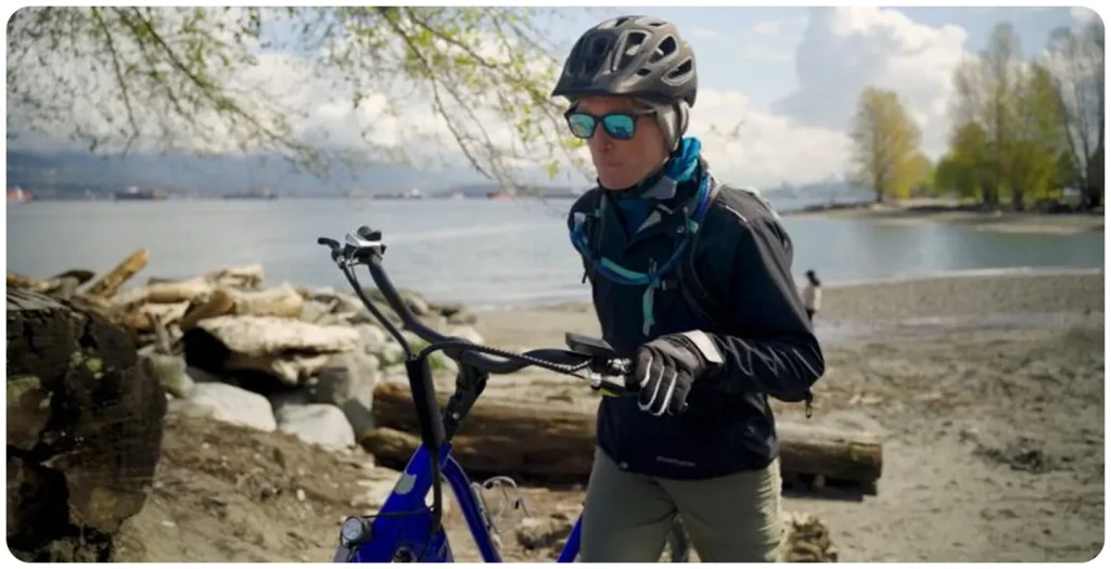 Cyclist wearing a helmet and sunglasses walking a blue bicycle along a rocky lakeside beach, with water and trees in the background on a clear day.