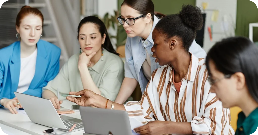 A diverse group of professional women collaborating around a table, reviewing information on laptops during a focused team meeting in a modern office environment.