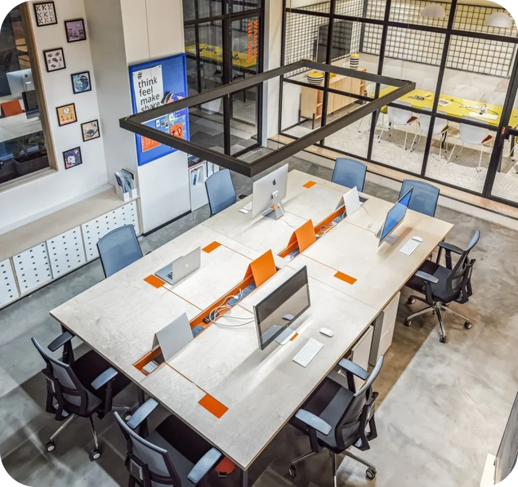 A modern open-plan office workspace with shared desks, desktop computers, ergonomic chairs, and glass-walled meeting rooms, viewed from above in a clean, minimalist design.